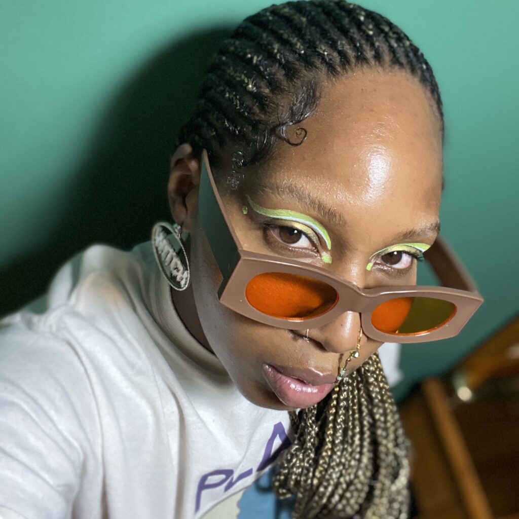 Photo of DJ Mystic Meg. She is making eye contact with the camera. Is wearing chunky brown sunglasses with orange lenses. She has braided hair and beautiful green and white eye makeup.