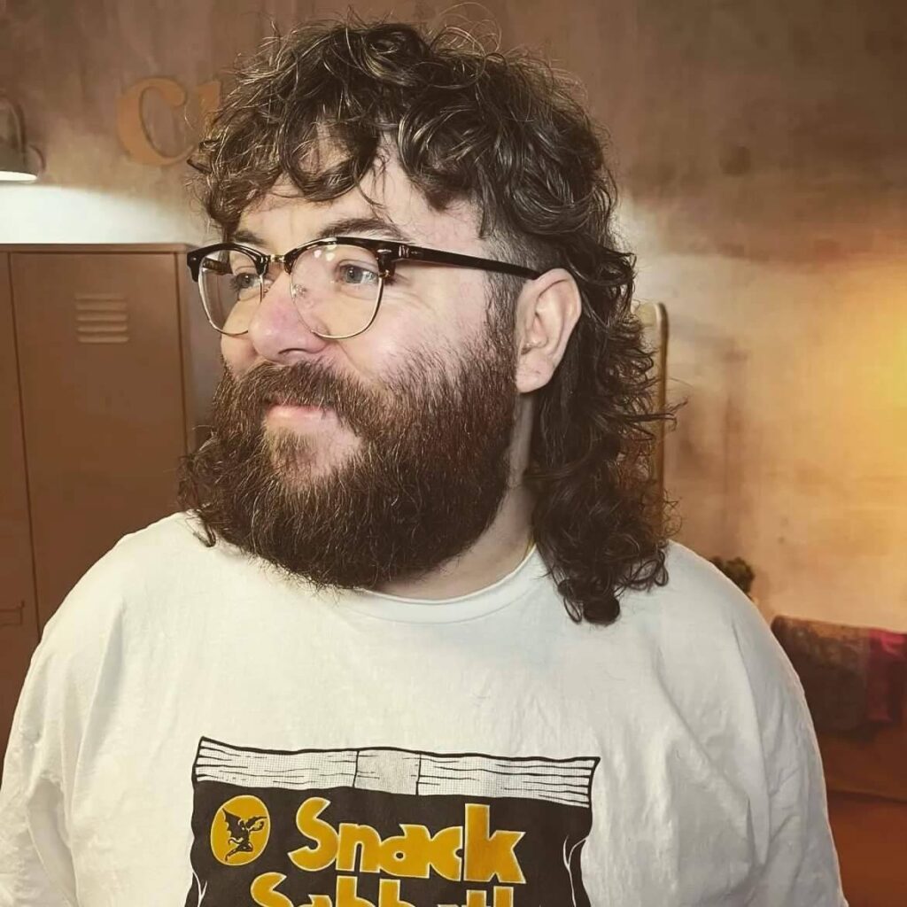 Photo of the DJ Scrapes. They are looking off camera to the side and smiling warmly. They have cool glasses and a brown curly beard and mullet.