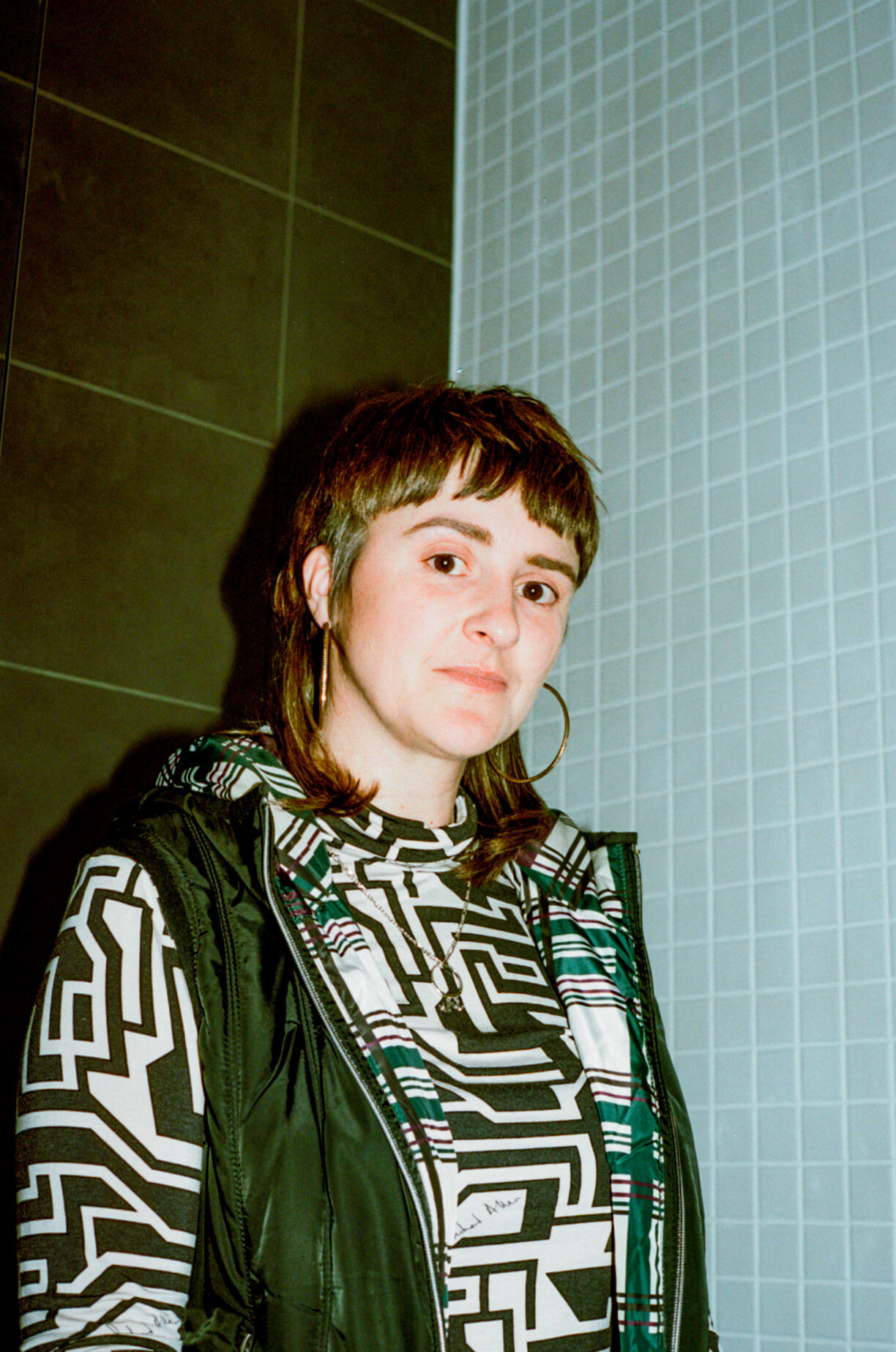 A photo of Sexy roy is looking at the camera wearing a funky black and white geometric top, hoop earrings. They are in a tiled room.