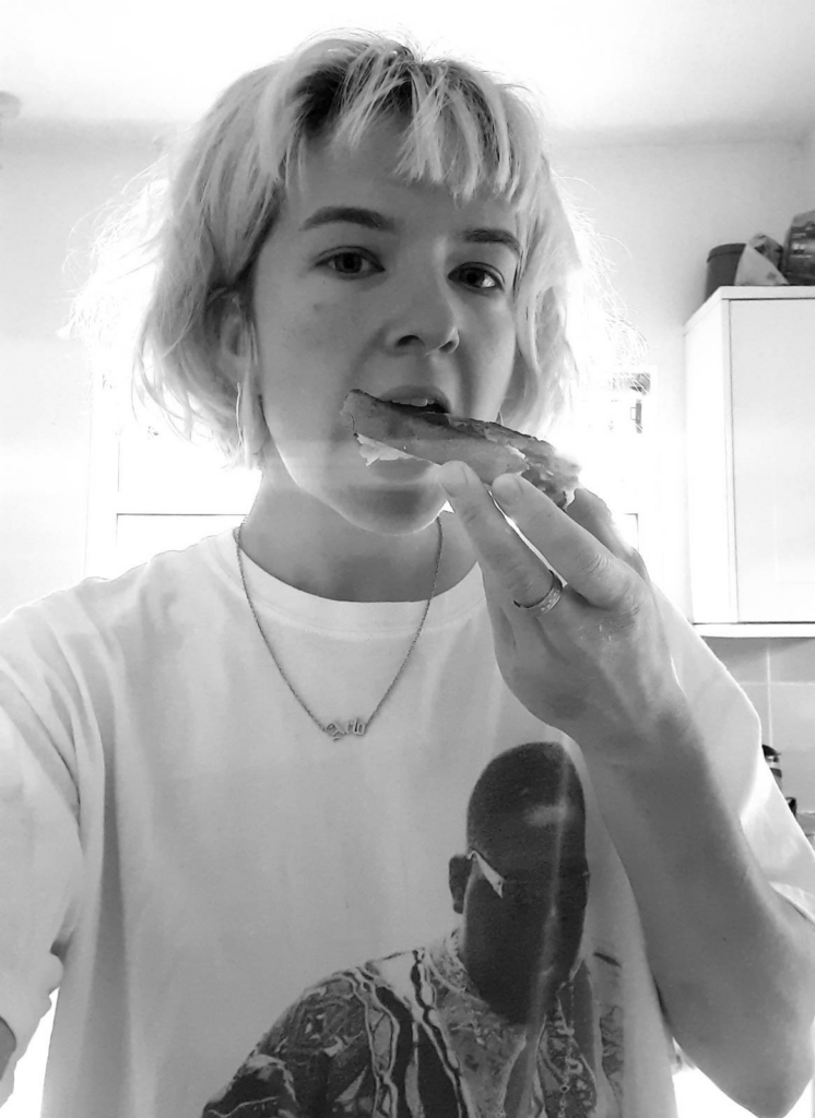 Photo of Arlo in black and white. They are in a kitchen wearing a white t-shirt looking at the camera whilst eating a piece of toast.