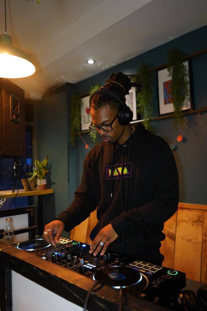 King Kai DJing at a party. He is wearing glasses, headphones and a black hoodie and multiple silver rings. His hair is tied up. He is DJing. There are plants and Keith Haring prints in the background.