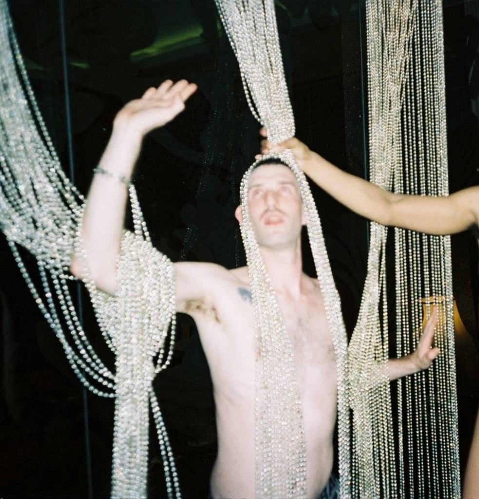 DJ Forgets is topless tangled up in a curtain of pearly beads. He is looking out the frame top left. A hand emerges from the right of the frame holding the beads over DJ Forget's head like a wig.