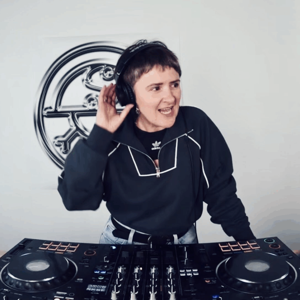 Roy behind the decks on Sigil Radio. In a black top and jeans. Their hand is up to their heapdhones and they are looking off screen.