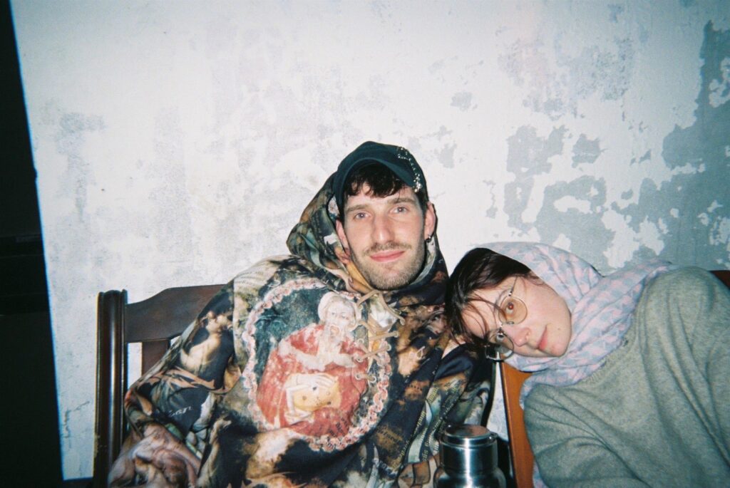 Analogue looking photo of DJ Forrgets. He is sitting in a chair in front of wall with paint peeling. He is wearing a cap and a hoodie with the hood up. The hoodie is very colorful print of some sort of old painting of tapestry. Another person called Stan is leaning into the photo onto DJ Forget's should. They are wearing glasses and a headscarf and a grey jumper.